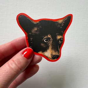 May include: A die-cut sticker featuring the head of a black and tan dog with pointed ears. The illustration uses detailed black linework and shading on a tan base, surrounded by a bold, solid red border that follows the contour of the dog's head.