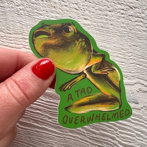 May include: A green sticker with a cartoon frog illustration and the words "A Tad Overwhelmed." The frog is in shades of green and brown. The sticker has a green border and is held by a person.