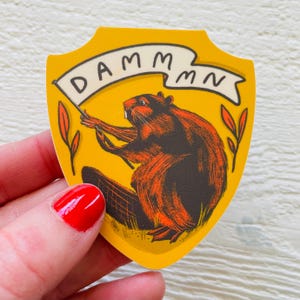 May include: A yellow shield-shaped sticker with a cartoon beaver holding a banner that reads "DAMMMN".
