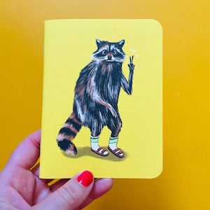 May include: A yellow notebook with a raccoon illustration. The raccoon is shown standing, wearing socks and sandals, and making a peace sign. The raccoon has brown and black fur and a striped tail.