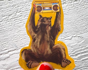 Pode incluir: Adesivo de um urso castanho com uma borda amarela, segurando um boombox vintage acima da cabeça. O boombox é amarelo, azul e castanho. O urso está sentado com as pernas cruzadas.