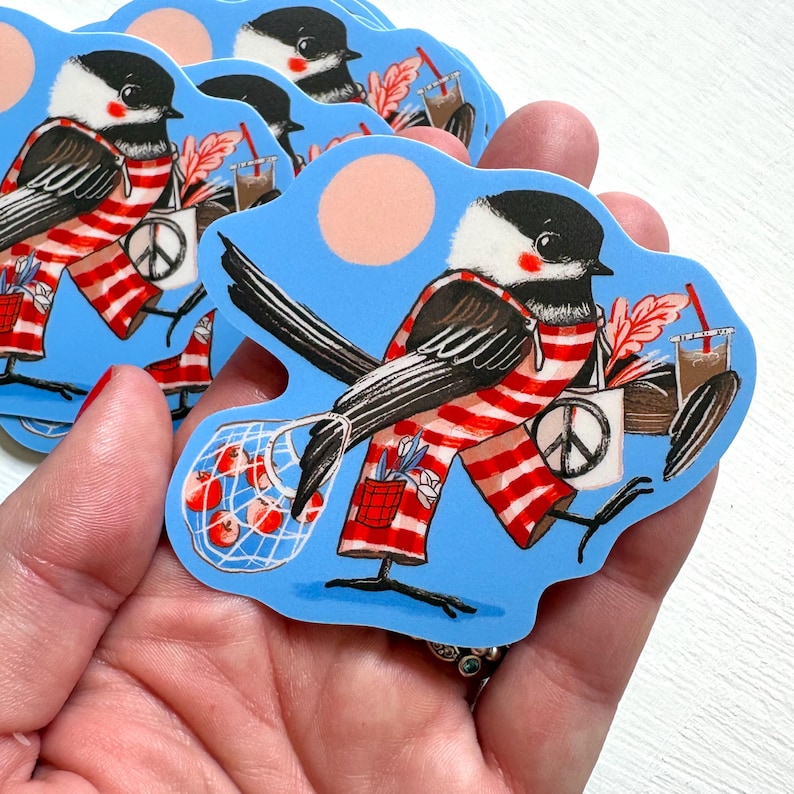 May include: A die-cut sticker featuring a black-capped chickadee wearing red and white striped overalls. The bird carries a mesh bag of red apples, a white tote bag with a peace sign, and a brown drink with a red straw against a blue background.