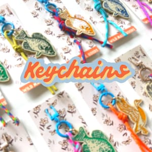 Peut inclure: Plusieurs porte-clés en forme de poisson aux couleurs et motifs variés. Les porte-clés sont attachés à des cordons colorés et à des fermoirs bleus. Le mot "Keychains" est écrit en orange et bleu sur l'image.