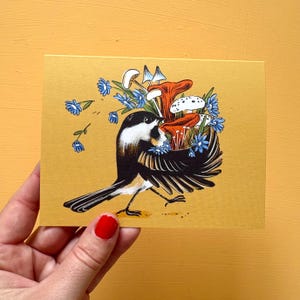 Peut inclure: Une carte de vœux rectangulaire sur fond jaune moutarde illustrant une mésange à tête noire. Le dos de l'oiseau est orné d'un amas de champignons orange, blancs et bleus, accompagnés de petites fleurs bleues dessinées sur le papier.