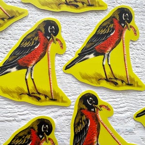 May include: Several yellow stickers featuring a stylized robin with a worm in its beak. The birds have black heads, wings, and tails, with red breasts and a long, thin worm. The stickers are on a white surface.