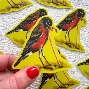 May include: A collection of yellow stickers featuring a stylized illustration of a robin with a worm in its beak. The bird has a red breast, black wings, and a black head. The stickers are on a white surface.
