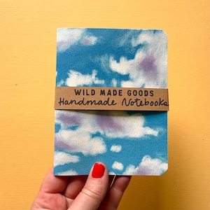 May include: A rectangular notebook featuring a blue sky with white and purple clouds printed on the cover. A brown paper band wraps around the centre with the text WILD MADE GOODS Handmade Notebooks printed in black ink on the front surface.