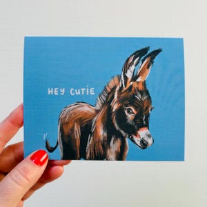 May include: A greeting card with a blue background featuring a brown and black donkey illustration. The text "HEY CUTIE" is written in white. The donkey has long ears and a white muzzle.