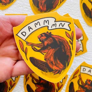 May include: A yellow shield-shaped sticker with a cartoon beaver holding a banner that reads "DAMMMMN". The beaver is brown and orange with black eyes and a white nose. The background is yellow with green leaves.