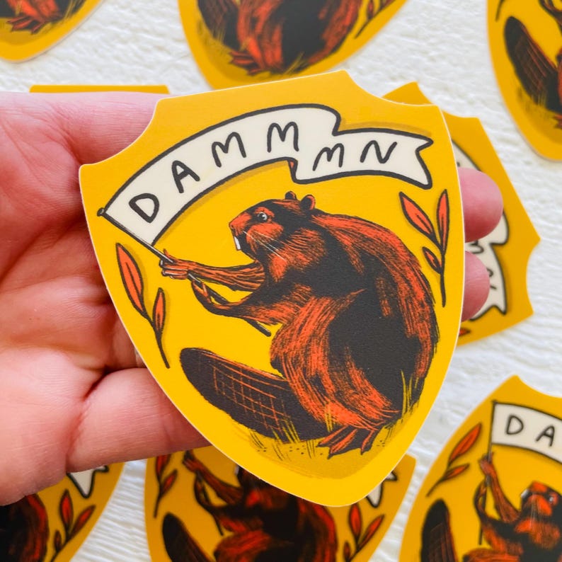 May include: A yellow shield-shaped sticker with a cartoon beaver holding a banner that reads "DAMMMMN". The beaver is brown and orange with black eyes and a white nose. The background is yellow with green leaves.