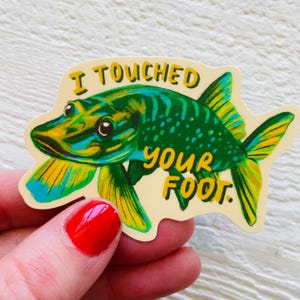 May include: A green and yellow fish sticker with the text "I touched your foot." The fish has a large mouth and a wide, flat body. The sticker is on a white background.