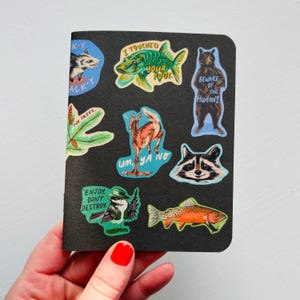 May include: A black notebook adorned with vibrant stickers depicting animals and text. Stickers showcase a possum, fish, bear, raccoon, deer, and bird. Phrases include "I touched your foot" and "Beware of the humans."