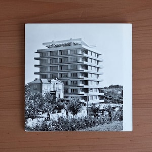 May include: Black and white photograph of a multi-story building with a unique architectural design. The building has multiple levels with horizontal lines and a flat roof. The image is on a white square card, set against a wooden background.