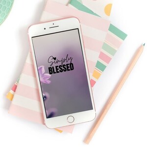 Puede incluir: Un teléfono inteligente blanco con una pantalla negra que muestra el texto "Simply Blessed" con un pequeño símbolo de corazón. La pantalla también muestra una flor morada. El teléfono está sobre una pila de papel de colores con patrones.
