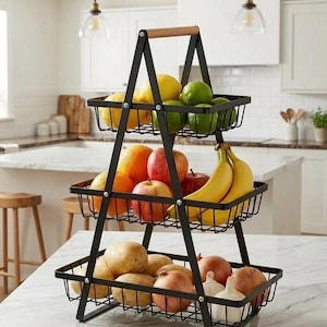 Könnte beinhalten: Ein dreistufiger Obstkorb aus schwarzem Metall mit Holzgriff, gefüllt mit Zitronen, Limetten, Orangen, Äpfeln, Bananen, Kartoffeln, Zwiebeln und Knoblauch. Der Korb steht auf einer weißen Arbeitsplatte.