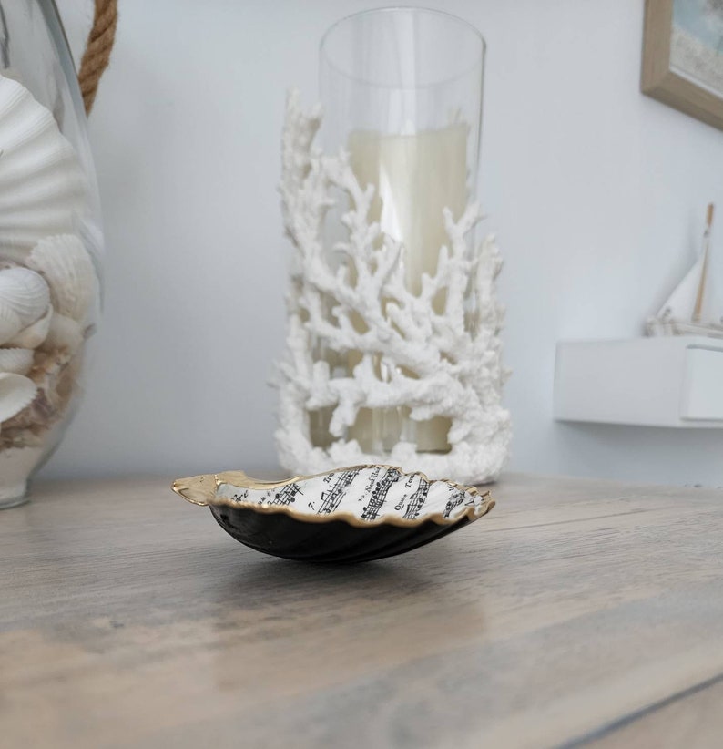 May include: A black and gold seashell dish with music notes printed on the inside. The dish is sitting on a wooden surface.