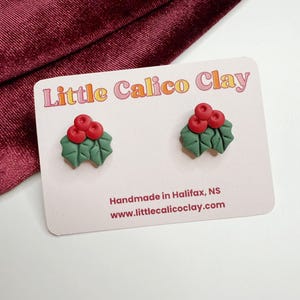 May include: Handmade holly stud earrings. Each earring has green leaves and three red berries. The earrings are presented on a card with the text "Little Calico Clay" and "Handmade in Halifax, NS".