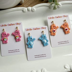 May include: A selection of bear-shaped earrings in pink, blue, and orange. The pink bears have rainbow details, the blue bears have a white belly, and the orange bears have a heart. Each pair is displayed on a card with the text "Little Calico Clay".