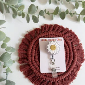 May include: A daisy-shaped badge reel with white petals and a yellow centre, attached to a clear retractable cord. The reel is on a burgundy macrame coaster, with the text "Little Calico Clay" on a white card.