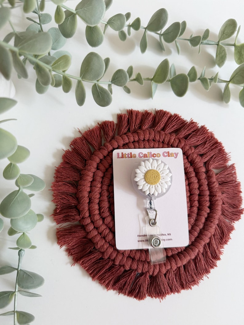 May include: A daisy-shaped badge reel with white petals and a yellow centre, attached to a clear retractable cord. The reel is on a burgundy macrame coaster, with the text "Little Calico Clay" on a white card.