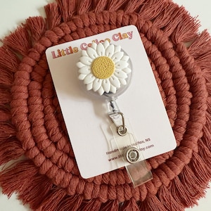 May include: A white daisy badge reel with a yellow centre is attached to a clear plastic retractable clip. The badge reel is on a white card with the text "Little Calico Clay". The background is a rust-coloured macrame coaster.