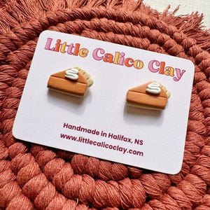 May include: Two handmade polymer clay earrings, each a slice of pumpkin pie with cream topping. The earrings are on a white card with the text "Little Calico Clay" and "Handmade in Halifax, NS". The background is a textured, rust-coloured surface.