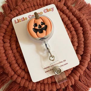 May include: A Halloween badge reel with an orange pumpkin design and a carved face. The reel is on a white card with the text "Little Coco Clay". The background is a rust-coloured macrame coaster.
