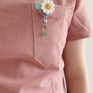 May include: A light pink scrub top with a pocket, featuring a daisy-shaped badge reel. The daisy has white petals and a yellow smiley face centre. A clear plastic ID holder hangs below the reel.