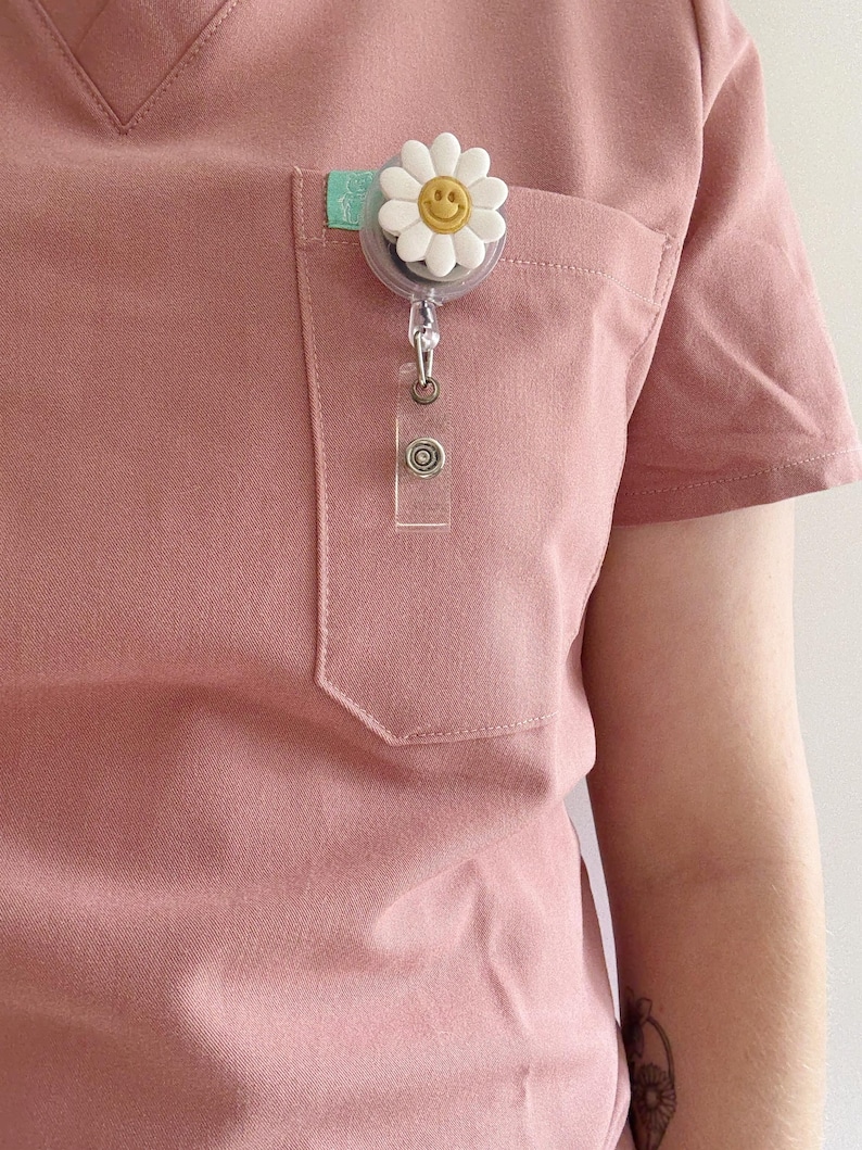 May include: A light pink scrub top with a pocket, featuring a daisy-shaped badge reel. The daisy has white petals and a yellow smiley face centre. A clear plastic ID holder hangs below the reel.