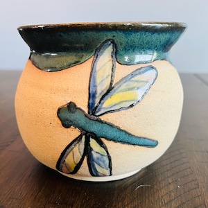 May include: A small, round, ceramic pot with a green glaze and a painted dragonfly design. The dragonfly has blue, yellow, and white wings and a green body.