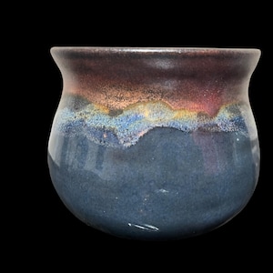 May include: A dark blue ceramic mug with a iridescent glaze. The glaze has a purple, pink, and blue color scheme.