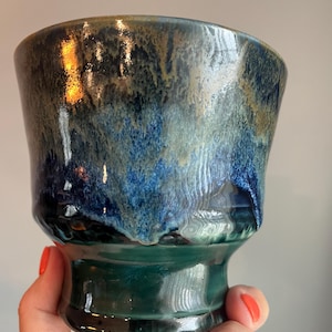 May include: Handmade ceramic cup with a unique glaze. The cup features a dark teal base and a gradient of blues and greens on the upper portion. The glaze has a glossy finish, adding depth and visual interest to the piece.