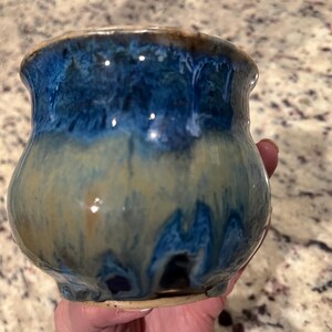May include: A handmade ceramic vase with a rounded body and a flared rim. The vase features a glossy glaze with a gradient of colors, transitioning from a deep blue at the top to a tan and cream color at the bottom. The vase is approximately 4 inches tall.