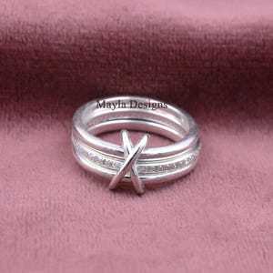 May include: A silver-toned ring featuring a central X-shaped crossover design and a band embedded with small clear stones. The ring is engraved with the text Mayla Designs on the inner surface, showcasing a polished metallic finish.