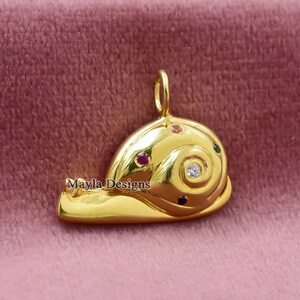 Könnte beinhalten: Goldfarbener Schneckenanhänger mit spiralförmigem Gehäuse. Das Gehäuse ist mit kleinen, farbigen Edelsteinen und einem zentralen, klaren Stein verziert. Der Anhänger hat eine Schlaufe für eine Kette und den Text "Mayla Designs".
