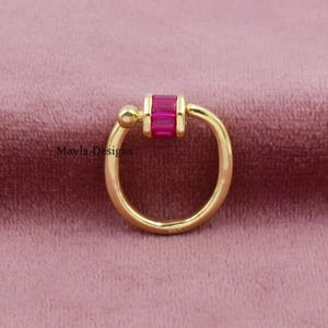 Puede incluir: Un anillo de aro dorado con una piedra rectangular de corte baguette en color rosa fucsia colocada horizontalmente en la parte superior. El anillo tiene un acabado liso y pulido, e incluye el texto Mayla Designs a la izquierda del aro.