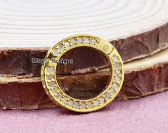 May include: A gold-colored, circular clasp with a ring of small, clear stones. The clasp is open, revealing a circular space in the center. The clasp is set on a wooden surface with a pink background. The text "Mayla Designs" is visible.