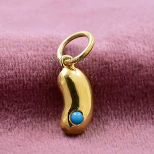 Puede incluir: Un dije dorado en forma de judía con una pequeña piedra redonda de color turquesa engastada en el centro. El dije tiene un lazo en la parte superior para sujetarlo a un collar o pulsera. El dije se muestra sobre un fondo rosa.