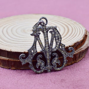 May include: A silver-toned pendant with the intertwined letters "ANB" encrusted with small, light-colored stones. The pendant has an ornate, vintage design and is displayed on a wooden surface against a pink background.
