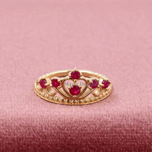 May include: A gold-coloured ring shaped like a crown, adorned with small, round, red gemstones. The ring sits on a pink surface. The text "Mayla Designs" is visible on the ring.