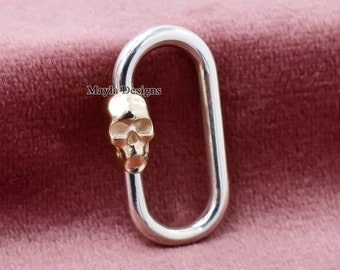 May include: A silver-colored oval-shaped clasp with a gold-colored skull accent. The skull is positioned on one end of the clasp. The clasp is displayed against a pink background. The text "Mayla Designs" is visible.