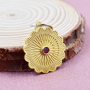 以下が含まれることがあります： 金色のペンダント。花柄のデザインで、中央に小さく丸い赤い宝石が配置されています。木製の背景とピンク色の表面に置かれています。