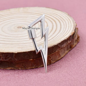 May include: A silver lightning bolt charm with a geometric design. The charm has a clasp at the top and a pointed bottom. The charm is displayed on a wooden slice with a pink background. The text "Mayla Designs" is visible.