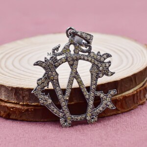 May include: A silver pendant with a monogram design featuring the letters 'W' and 'E' set with small diamonds. The pendant is on a wooden surface.