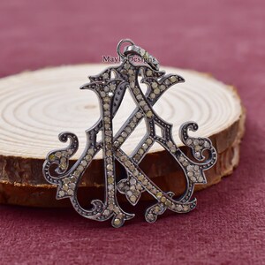 May include: A silver-toned pendant featuring an ornate intertwined "A" and "K" design, encrusted with small, light-colored gemstones. The pendant has a loop at the top for a chain and is set against a wooden surface.