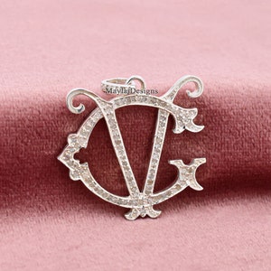 May include: A silver pendant with a diamond-encrusted monogram of the letters "CV". The pendant is on a pink velvet background. The text "Mayla Designs" is visible on the pendant.