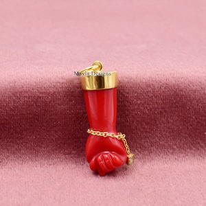 Puede incluir: Un colgante de puño de coral rojo con una cadena de oro y un pequeño acento de diamante. El colgante está sobre un fondo de terciopelo rosa. "Mayla Designs" está escrito en la parte superior del colgante.