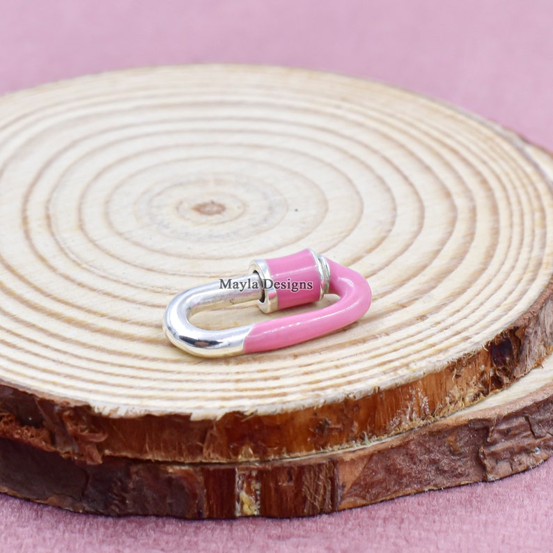 May include: A pink and silver metal clasp with a rounded shape. The clasp is on a wooden surface. Mayla Designs is written on the clasp.