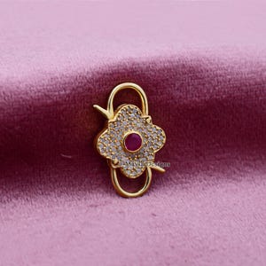 May include: A gold-colored jewelry clasp with a four-petal flower design. The center of the flower is a small, round, red gemstone, surrounded by smaller, clear gemstones. The clasp is on a pink background.