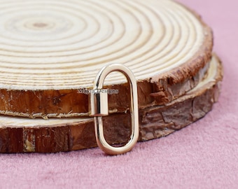 May include: A gold-colored oval-shaped clasp with a hinged closure. The clasp is displayed on a wooden slice with a pink background. The text "Mayla Designs" is visible on the wooden surface.